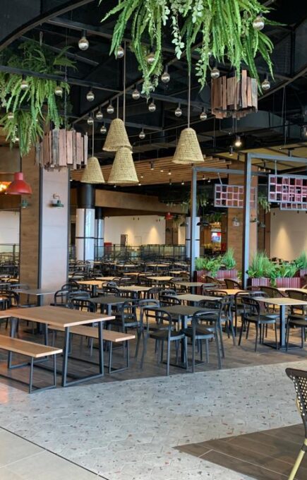 Área de comedor amplia y flexible, equipada con mesas grupales y bancas, ideal para food court o espacios de reunión. El diseño resalta por su iluminación cálida, abundante vegetación y detalles en madera, generando un ambiente acogedor y moderno.